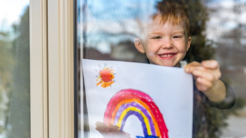 Young toddler smiling in a window showing a rainbow drawing