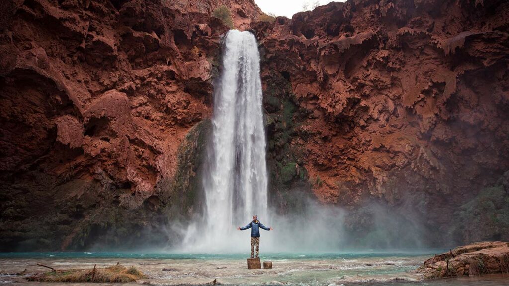 Neal Lee standing with arms outstretched in front of a majestic waterfall.