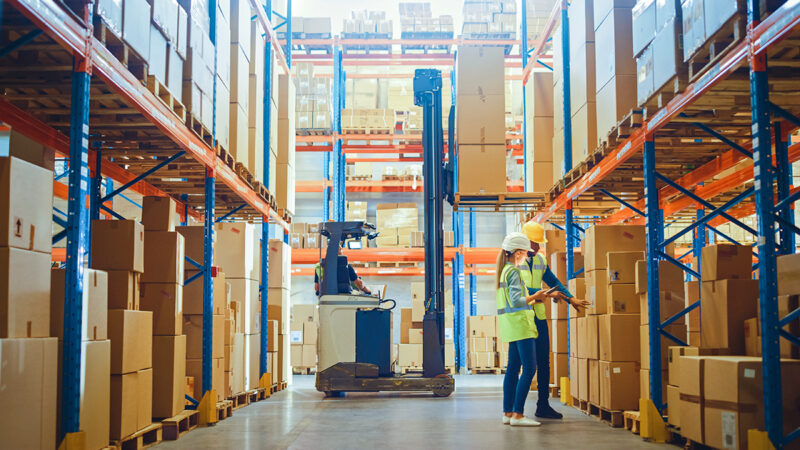 Retail Warehouse full of Shelves with Goods in Cardboard Boxes, Male and Female Supervisors Use Digital Tablet Discuss Product Delivery while Scanning Packages Forklift Working in Logistics Storehouse