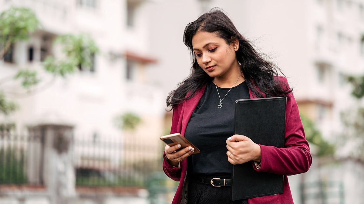 Business woman walking, holding a padfolio and looking at her smartphone.